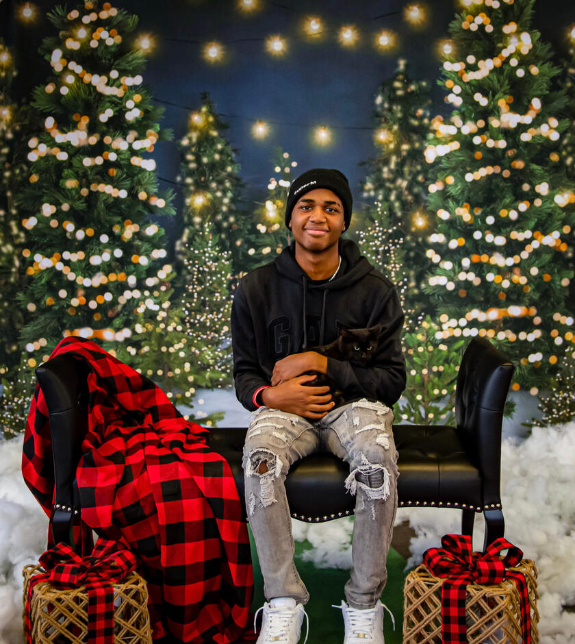 Maxwel, my younger brother, a black boy with rich brown skin, poses with a new kitten. The kitten, is small and black and nestled in his arms, looking away from the camera. There are premade Christmas photoshoot decorations in the background.